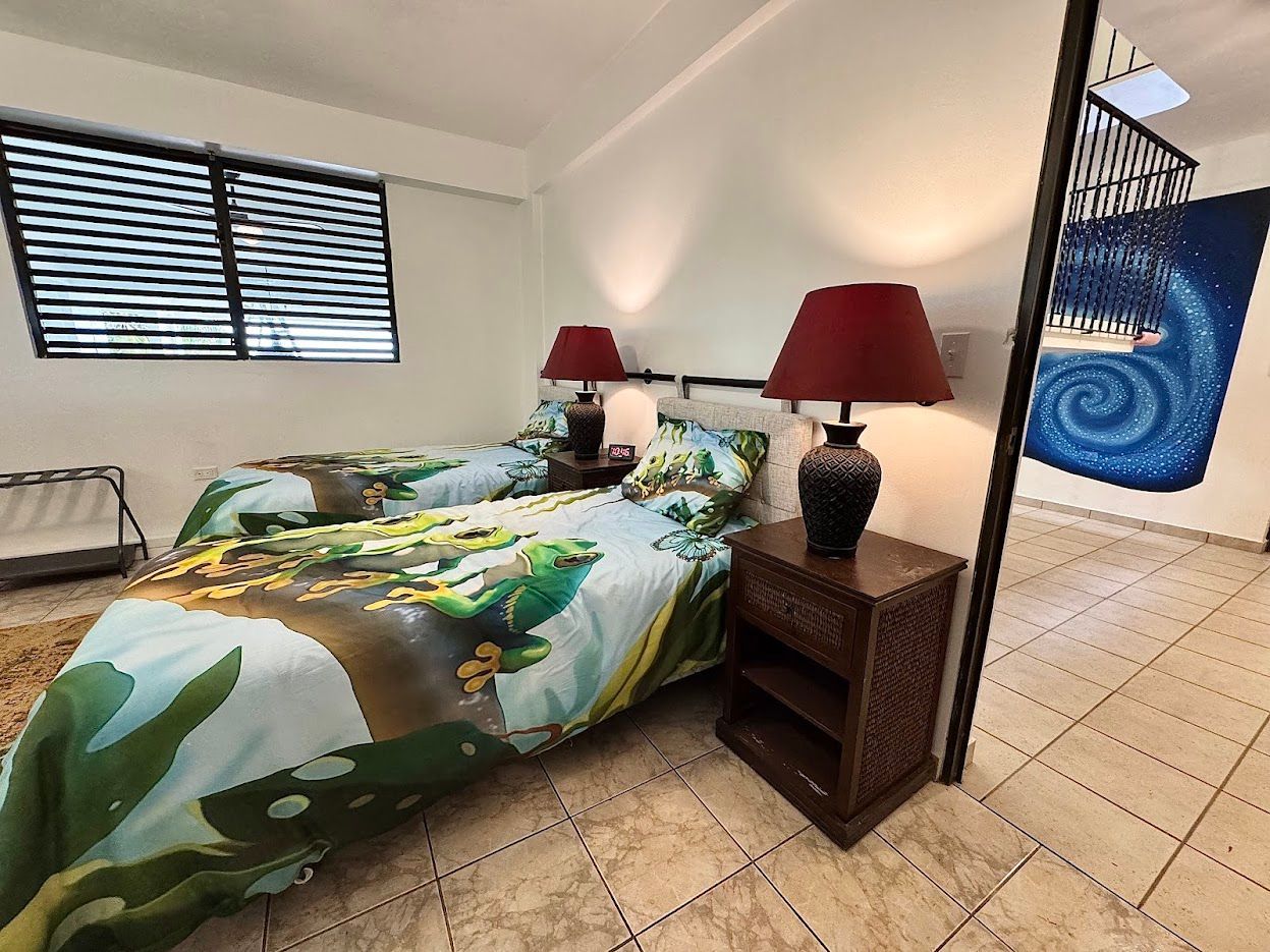 Twin beds with colorful bedding, lamps on a nightstand, and a window in a bedroom.