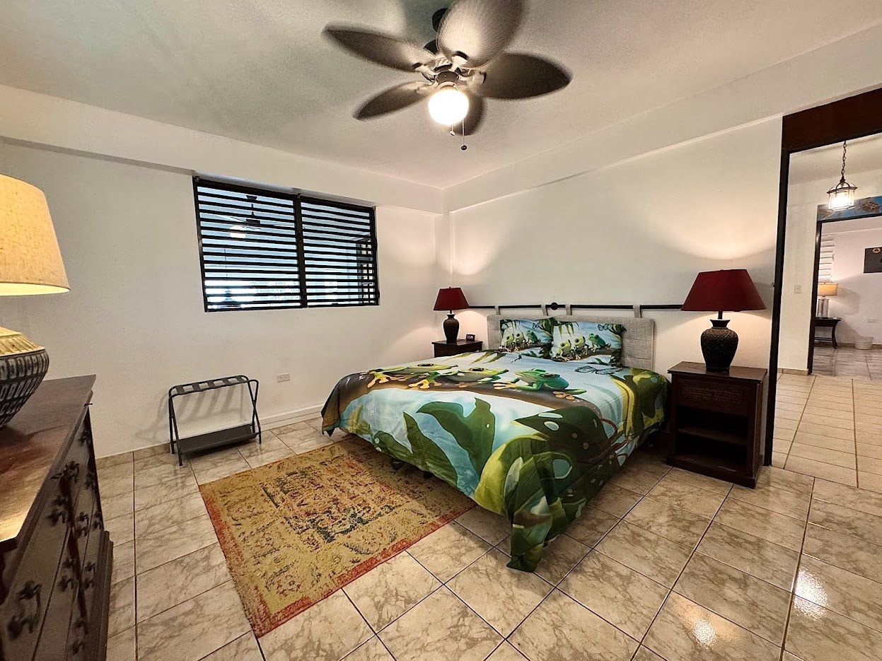 Bedroom with bed, nightstands, rug, and window with blinds. Ceiling fan and lamps.