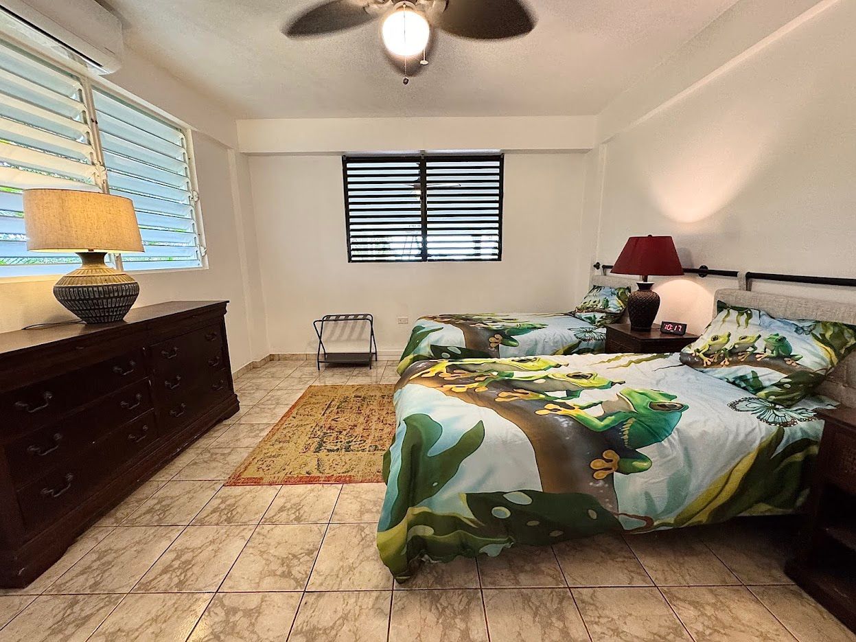 Bedroom with two beds, dresser, and windows with blinds. Earth tones, patterned bedding, and lamps.