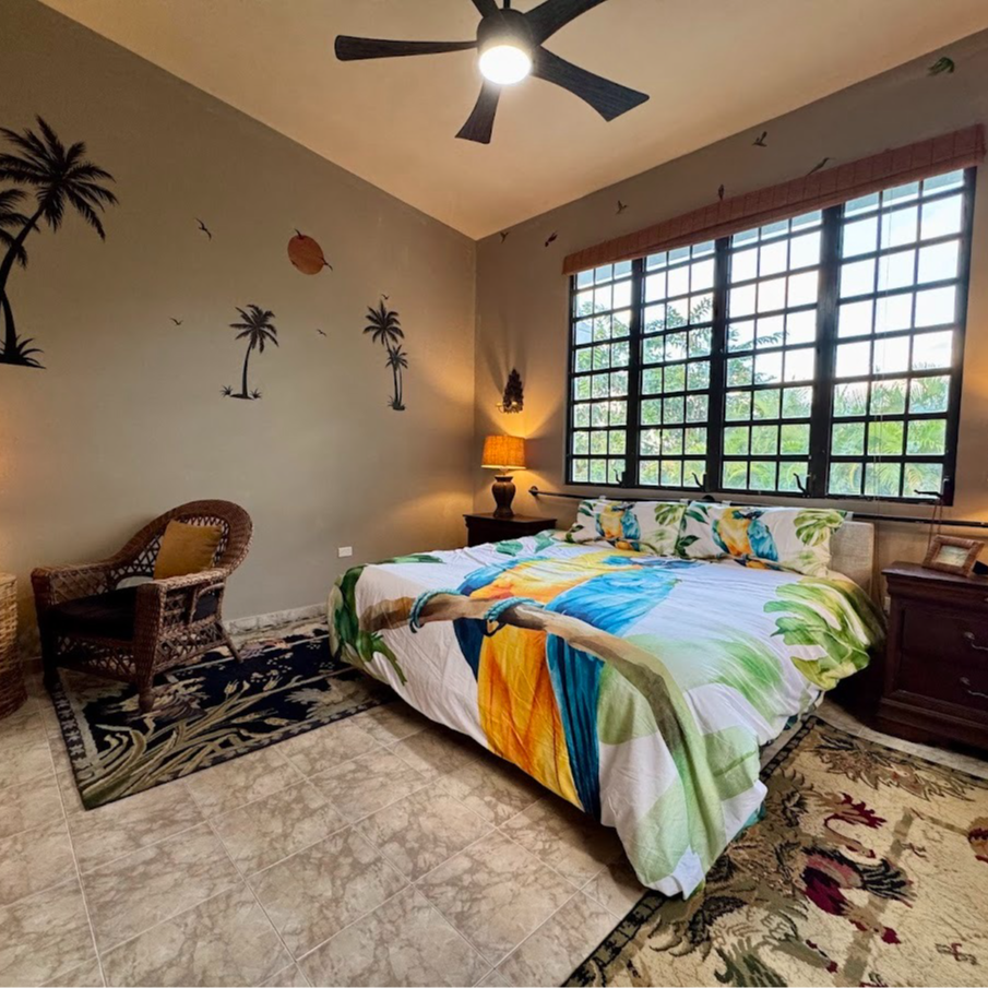 Bedroom with palm tree decor, bed with colorful linens, wicker chair, and large window.