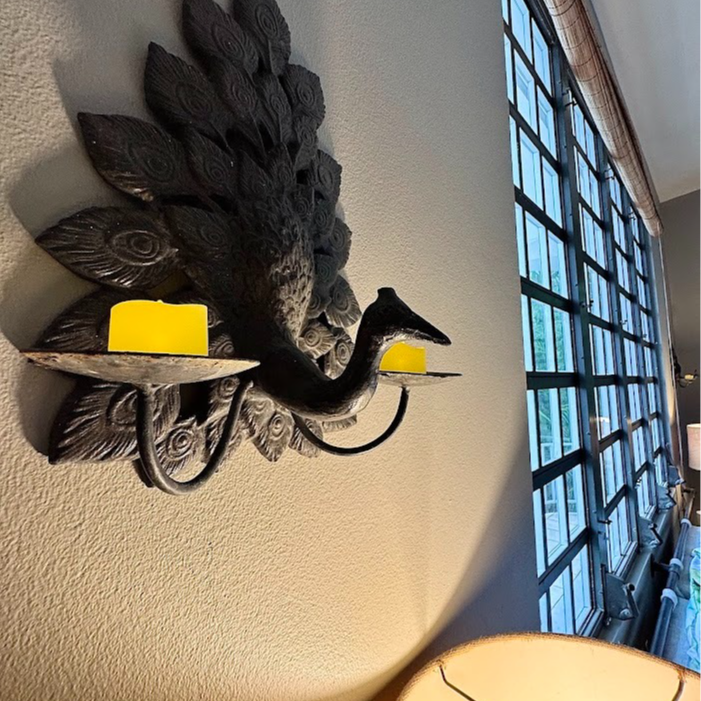 Black peacock wall sconce with yellow candles against a textured wall, next to a large window.