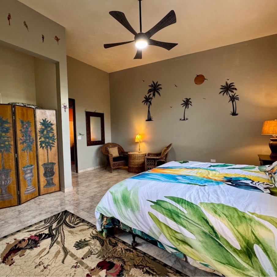 Bedroom with tropical theme; bed with botanical print, palm tree wall decals, wicker chairs, and folding screen.