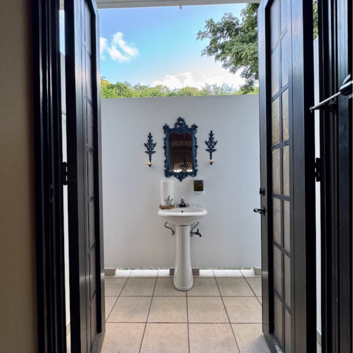 Open doorway leads to an outdoor bathroom: pedestal sink, ornate mirror, and blue sky.