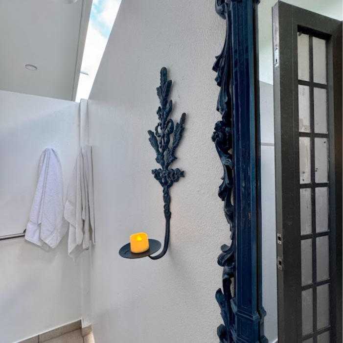 Black wall sconce with lit yellow candle on white wall, next to ornate dark blue object, towels in the background.