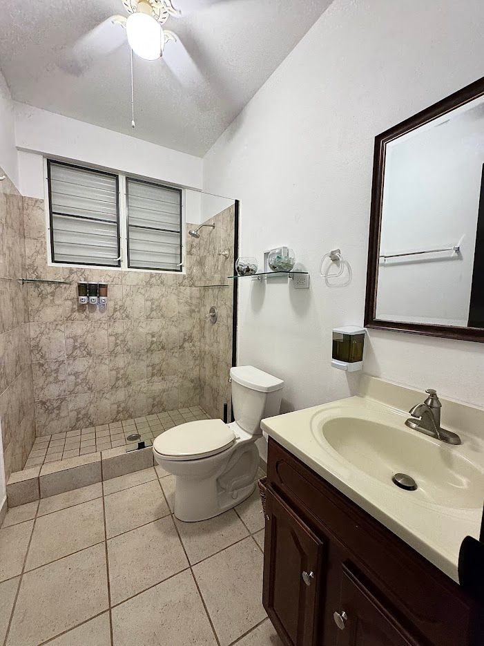 Bathroom with shower, toilet, vanity, and mirror. White walls, tile floors, and a ceiling fan.