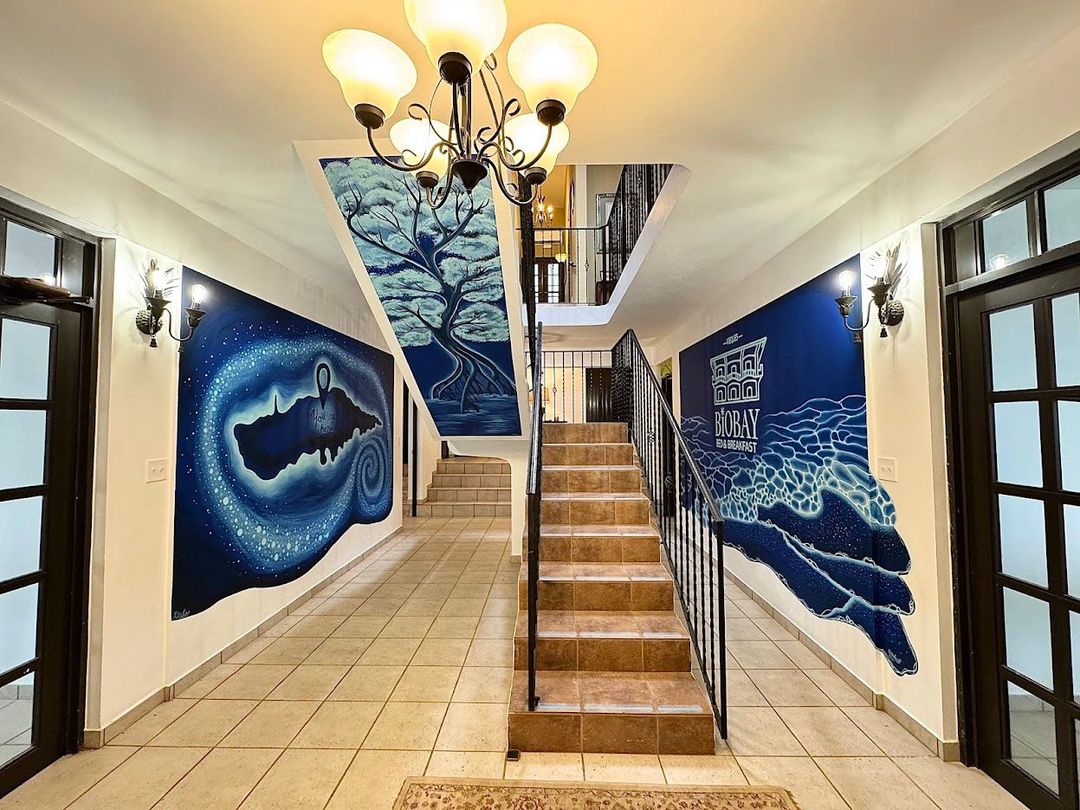 Hallway with staircase, murals, and chandelier. Blue and white artwork adorns walls, featuring nature scenes.
