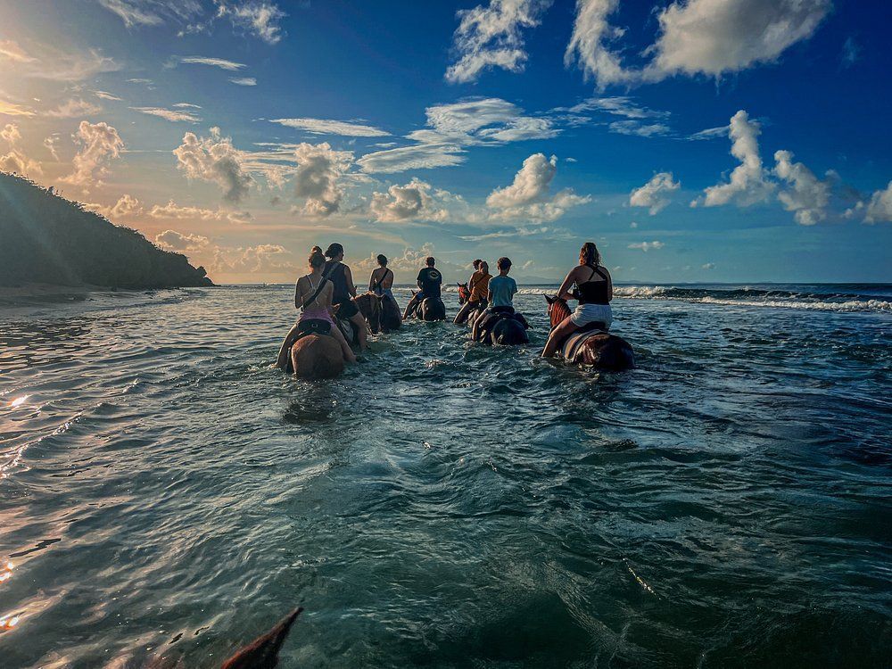 People on horseback ride through ocean water, blue sky and clouds.