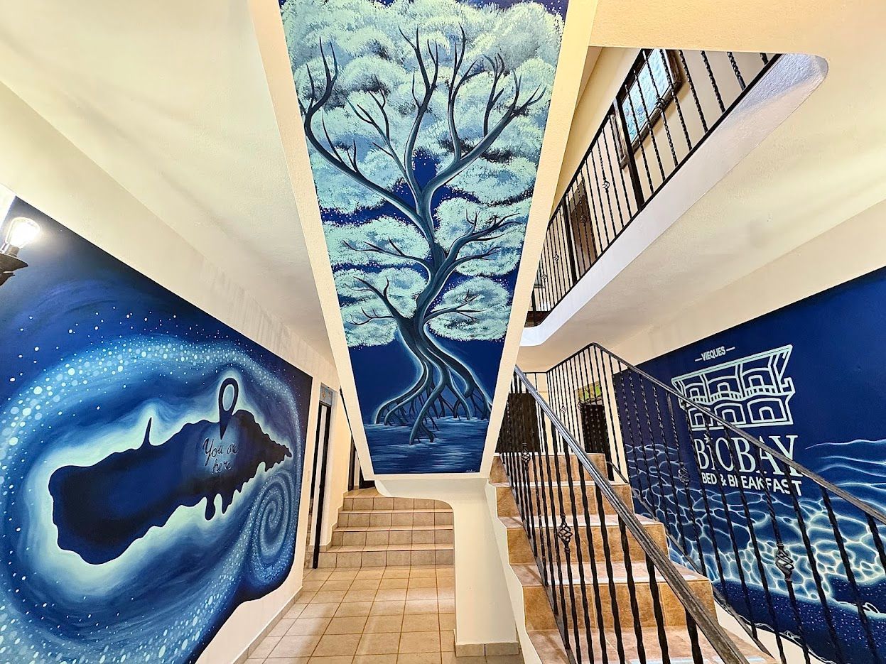 Blue-painted hallway with staircase; tree mural on ceiling, other murals on side walls.