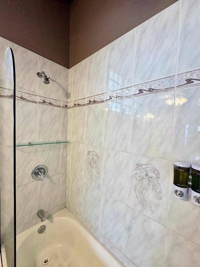 Tiled bathroom with a bathtub, showerhead, glass shelf, and mounted soap dispensers. Brown accent on top.