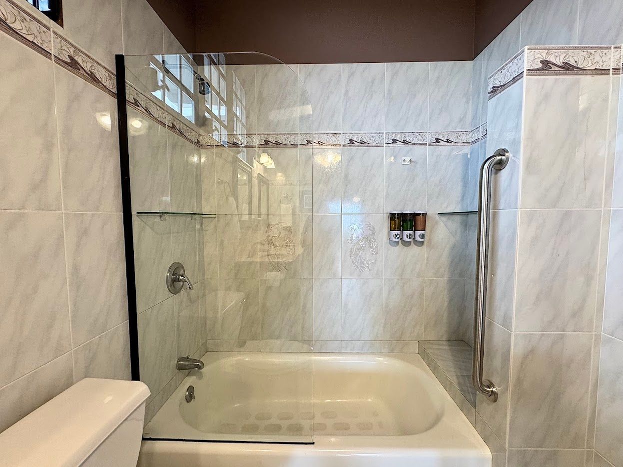 Bathroom with white tub, glass shower door, and grab bar. White tile walls and brown trim.