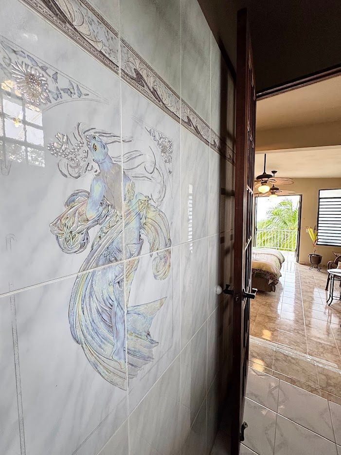 Tiled wall with mermaid art, ajar wooden door, and a glimpse into a room with bed and fan.