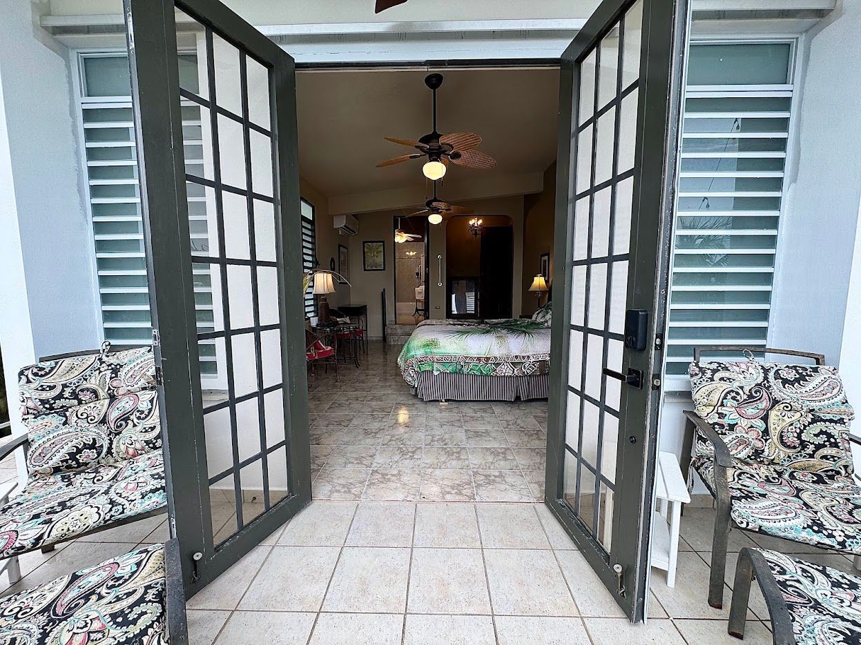 Open French doors reveal a bedroom with a bed. Two chairs sit on the balcony outside.