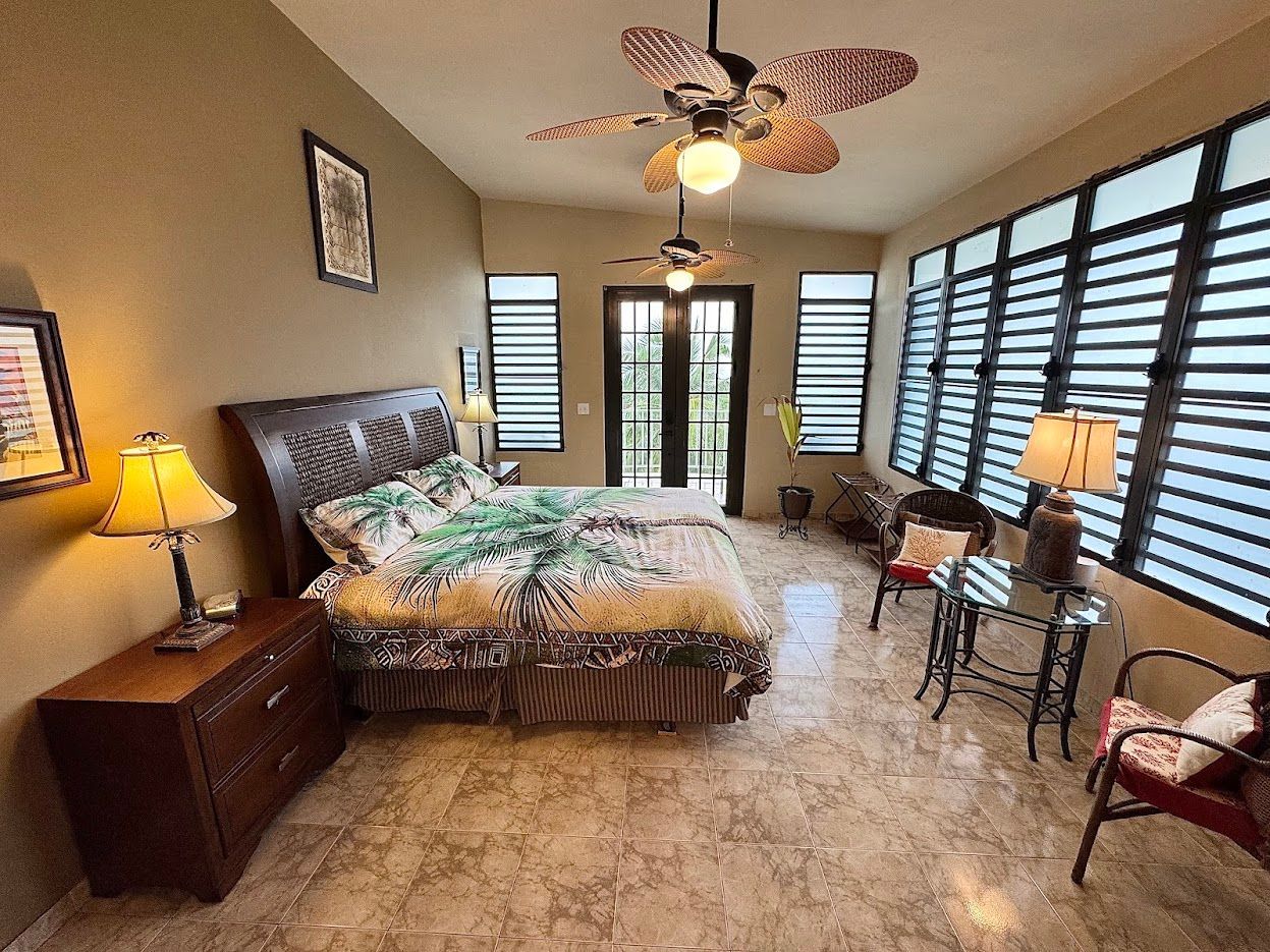 Bedroom with bed, nightstands, ceiling fans, shuttered windows, and sitting area.