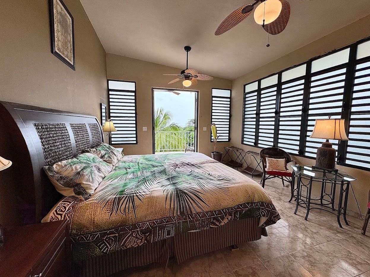 Bedroom with bed, side table, windows with horizontal blinds, open doorway.