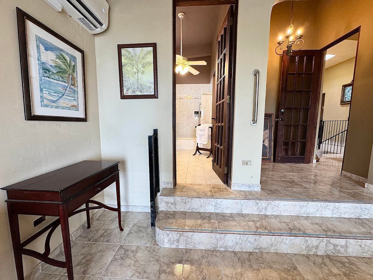 Entryway with steps, console table, artwork, open doors to rooms with tile floors and chandeliers.