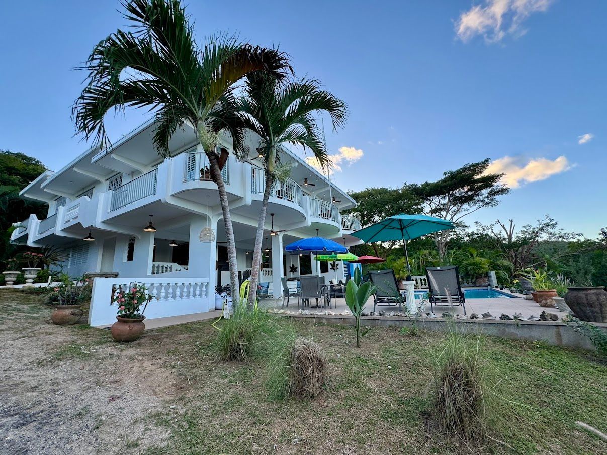 White multi-story building with palm trees, outdoor seating, and umbrellas against a blue sky.