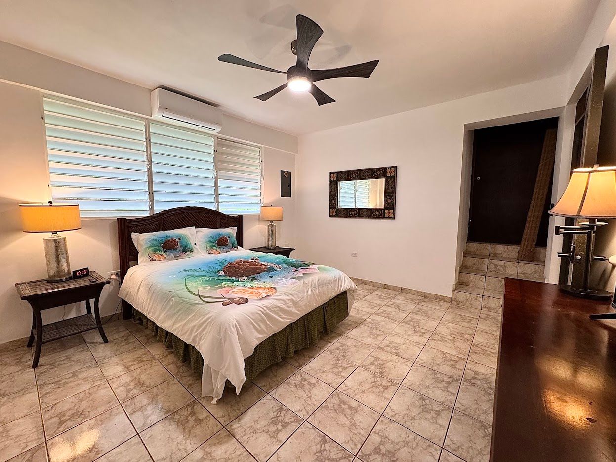 Bedroom with a bed, nightstands, lamps, ceiling fan, blinds, and a door.