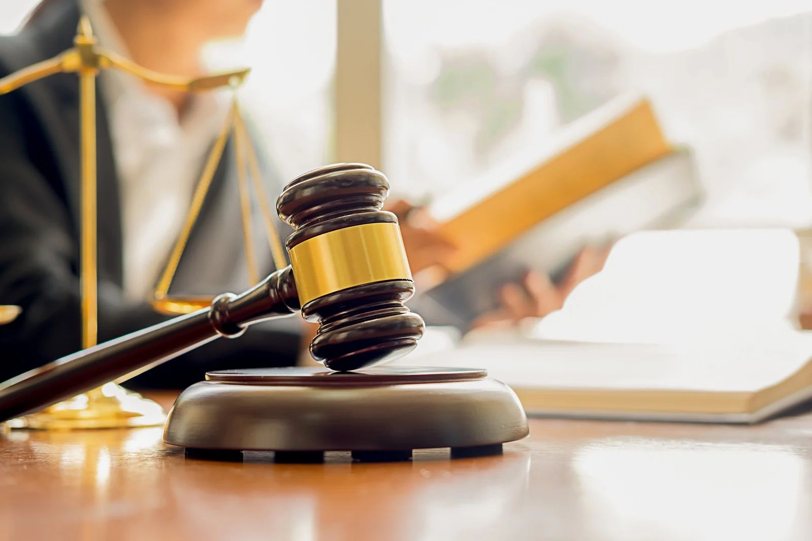 Gavel on a wooden desk with a person reading law books and a scale of justice in the background.