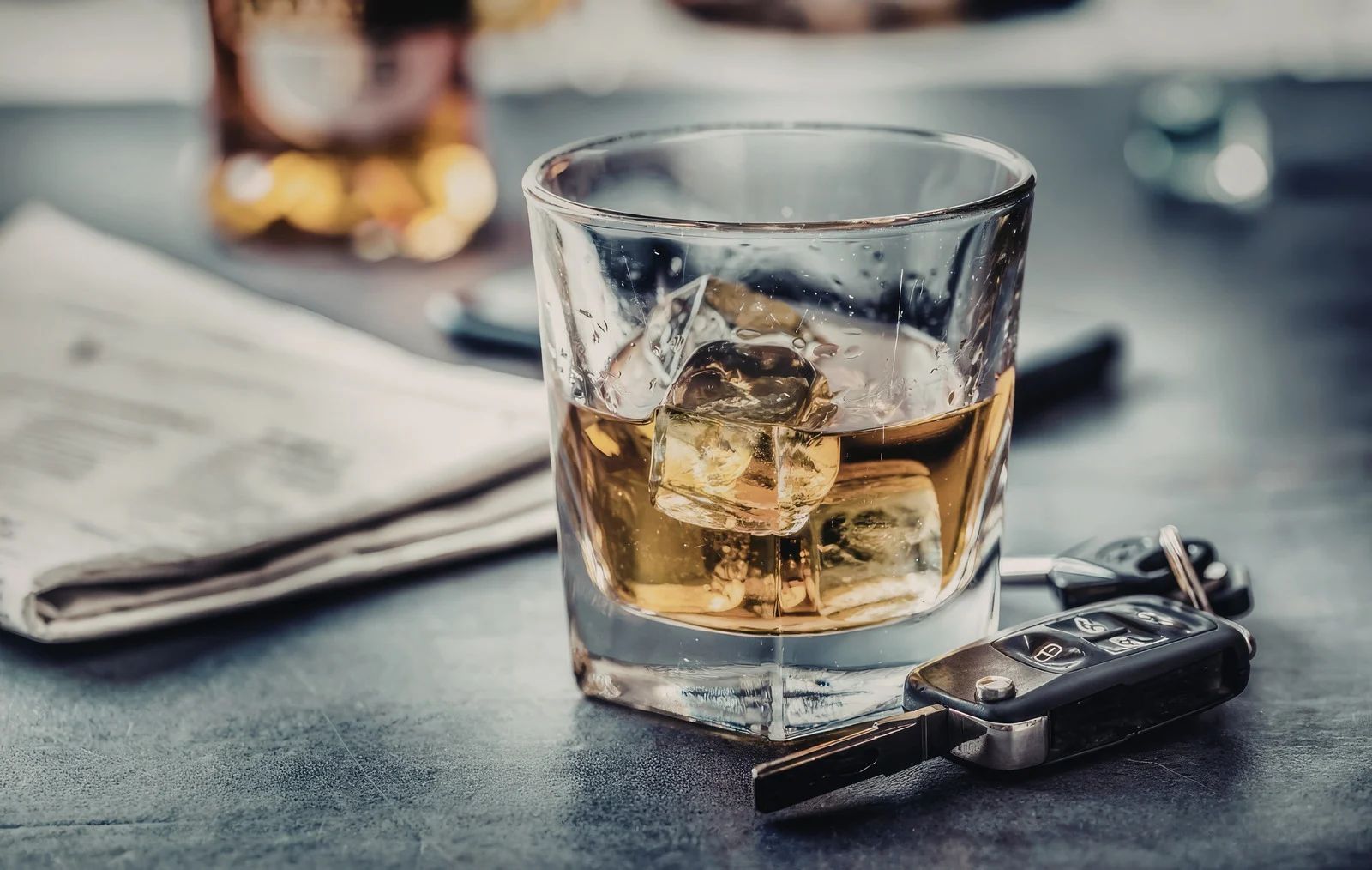 Whiskey glass with ice on a bar beside a set of keys and folded newspaper