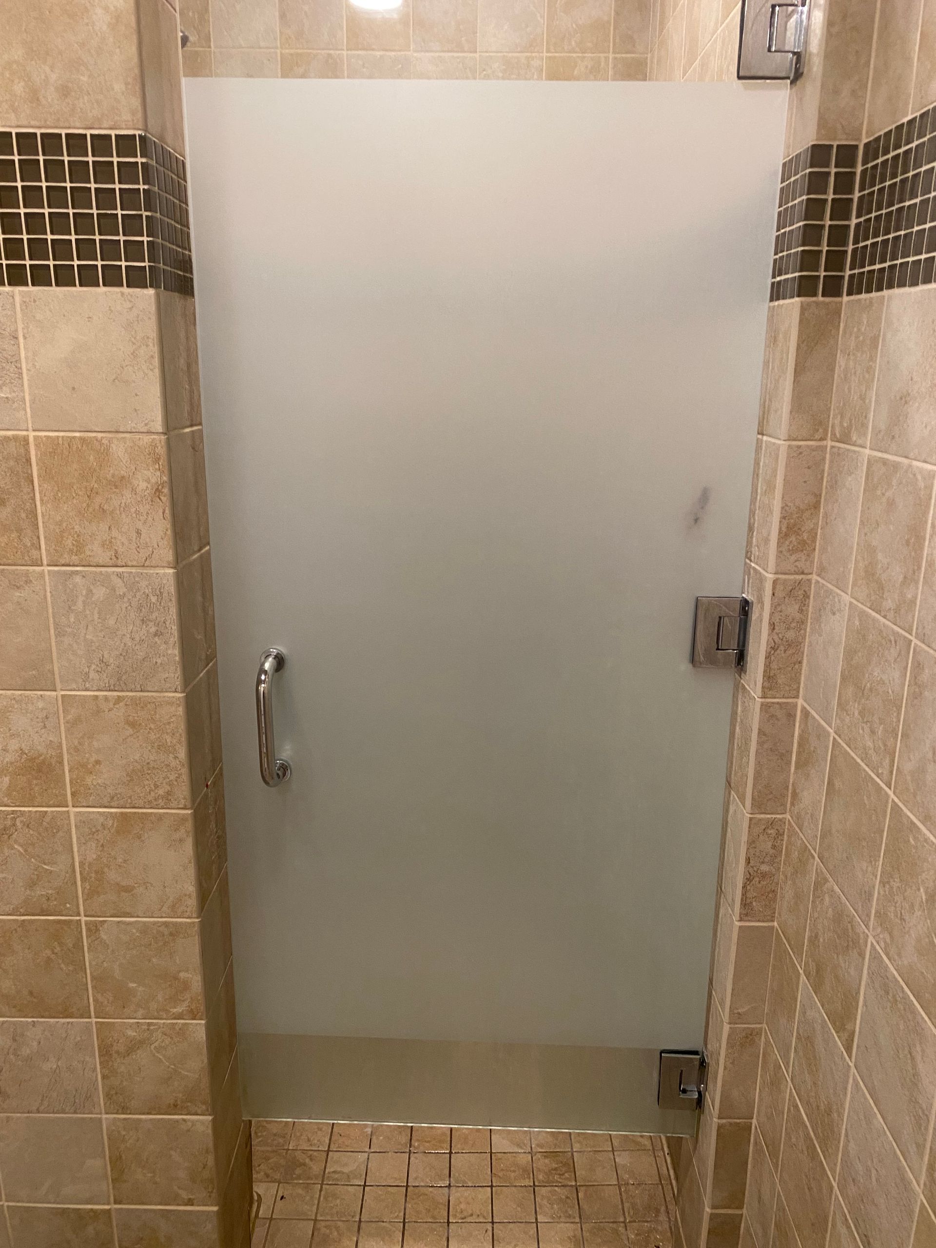 Frosted glass bathroom stall door with a silver handle and metal latch, set between beige tiled walls.