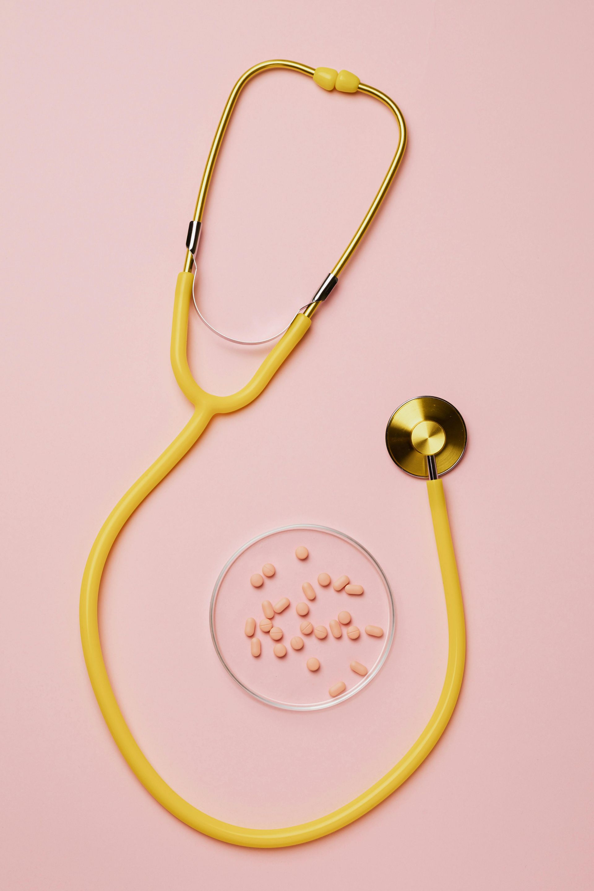 Yellow stethoscope with a gold chestpiece on a pink background beside a petri dish of pills