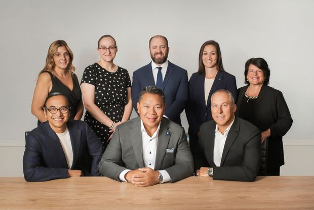 Group of nine people posing in front of a wooden table and a white background. Men and women wearing professional attire.