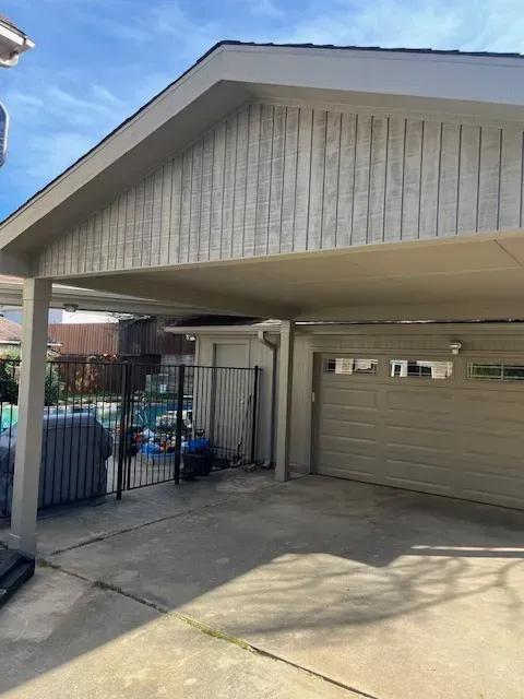 Carport with a beige garage door, concrete driveway, and a fenced storage area under a gray roof.