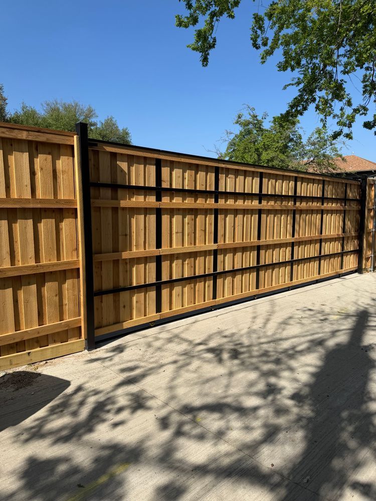 Wooden privacy fence along a driveway under a clear blue sky, with tree shadows on the pavement.