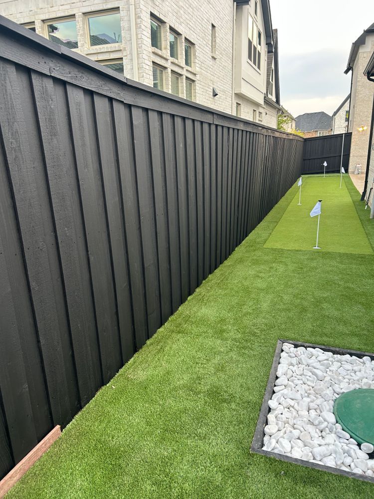 Narrow artificial turf side yard with black fence and white stone border alongside modern buildings