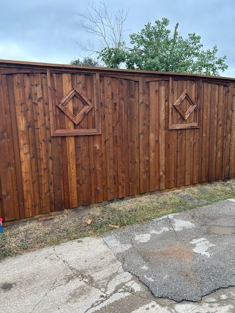 Wooden privacy fence with decorative diamond accents beside a cracked paved driveway and trees behind it