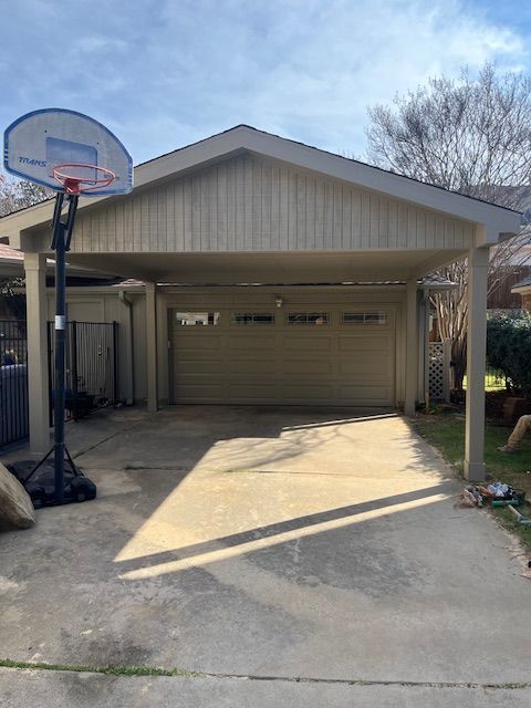 Carport with a closed garage door and a basketball hoop on the left, in bright daylight.