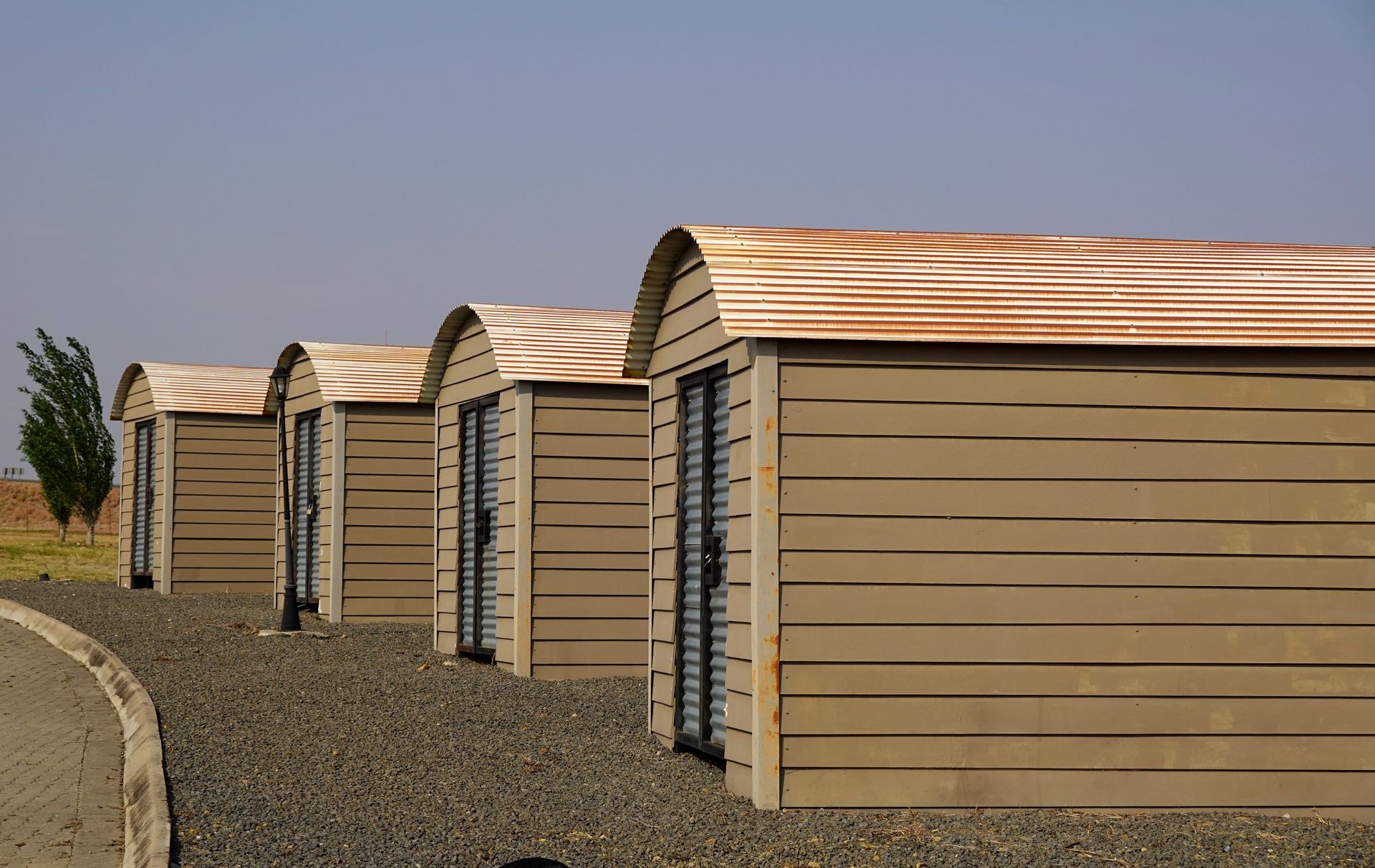 Row of small beige sheds with rusty metal roofs on a gravel lot
