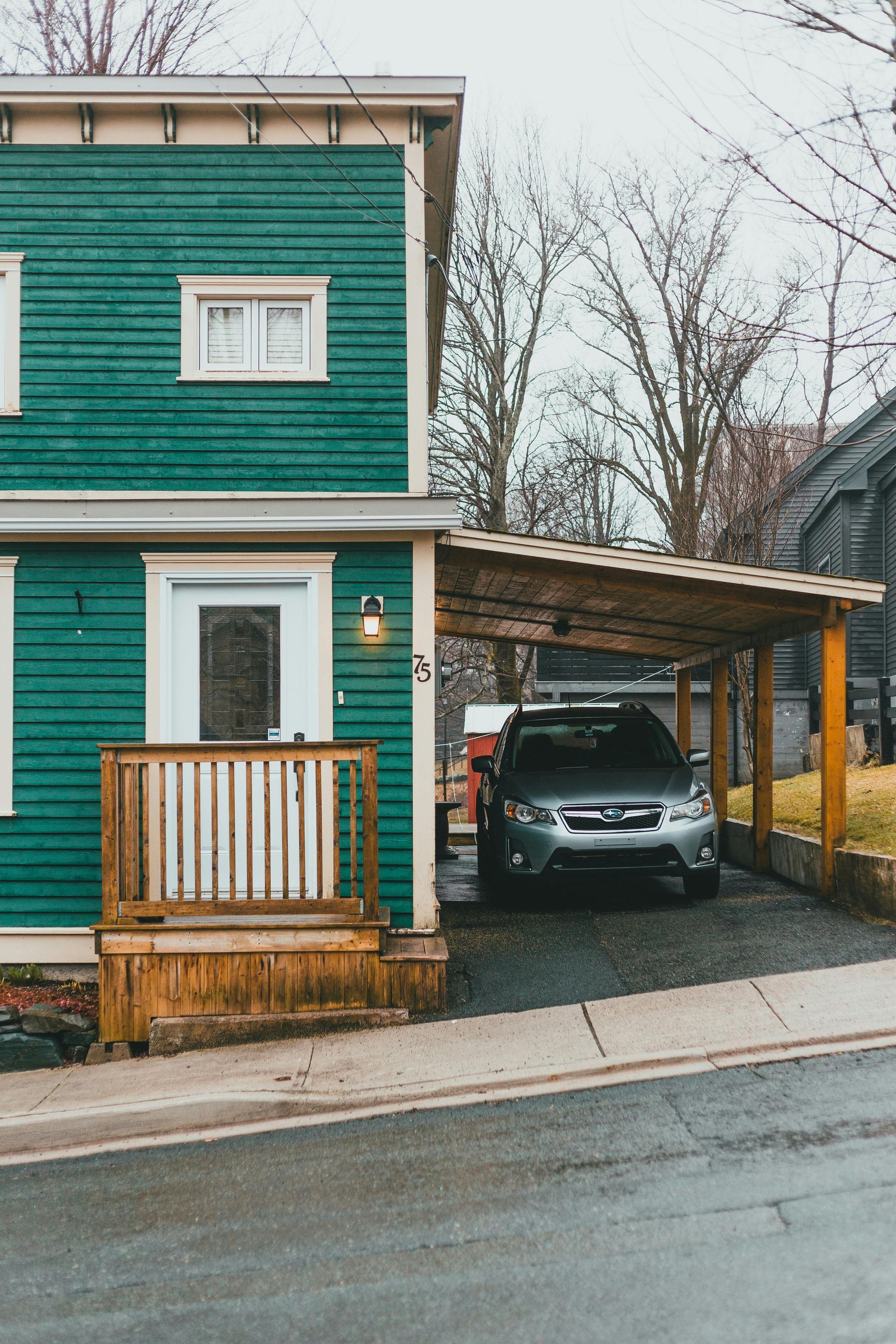 Green house with white trim and a car parked under a wooden carport beside the street