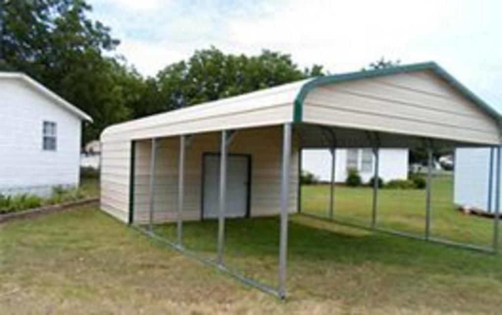 A carport with a green roof is sitting in the grass in front of a house.