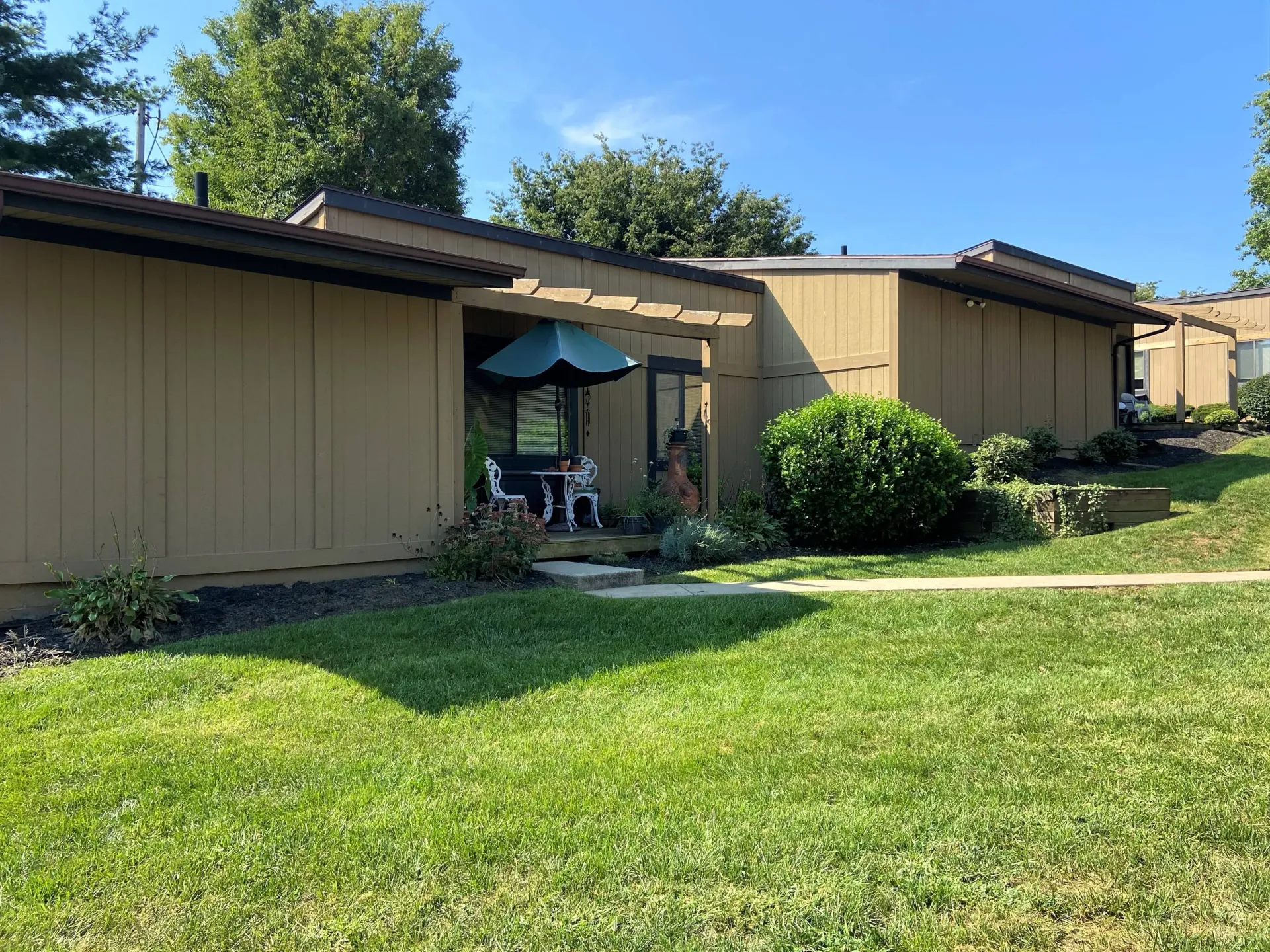 Tan townhouses with brown trim and awnings; sunny day with green grass and shrubs.