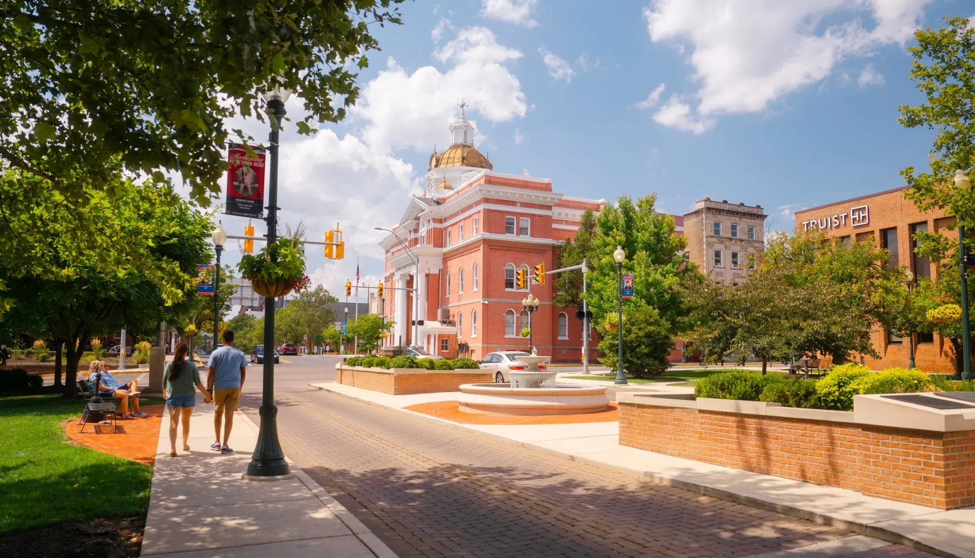 Couple walks down a brick street towards a historic brick building with a gold dome, trees, and gardens.