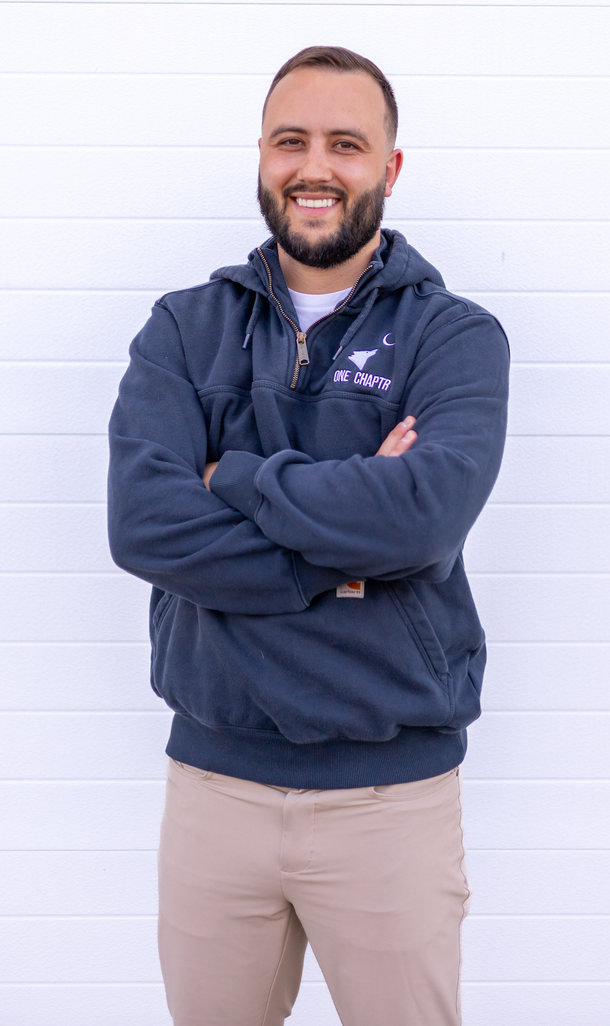 Man with beard, arms crossed, smiling. Wearing navy blue hoodie, tan pants, standing in front of a white wall.