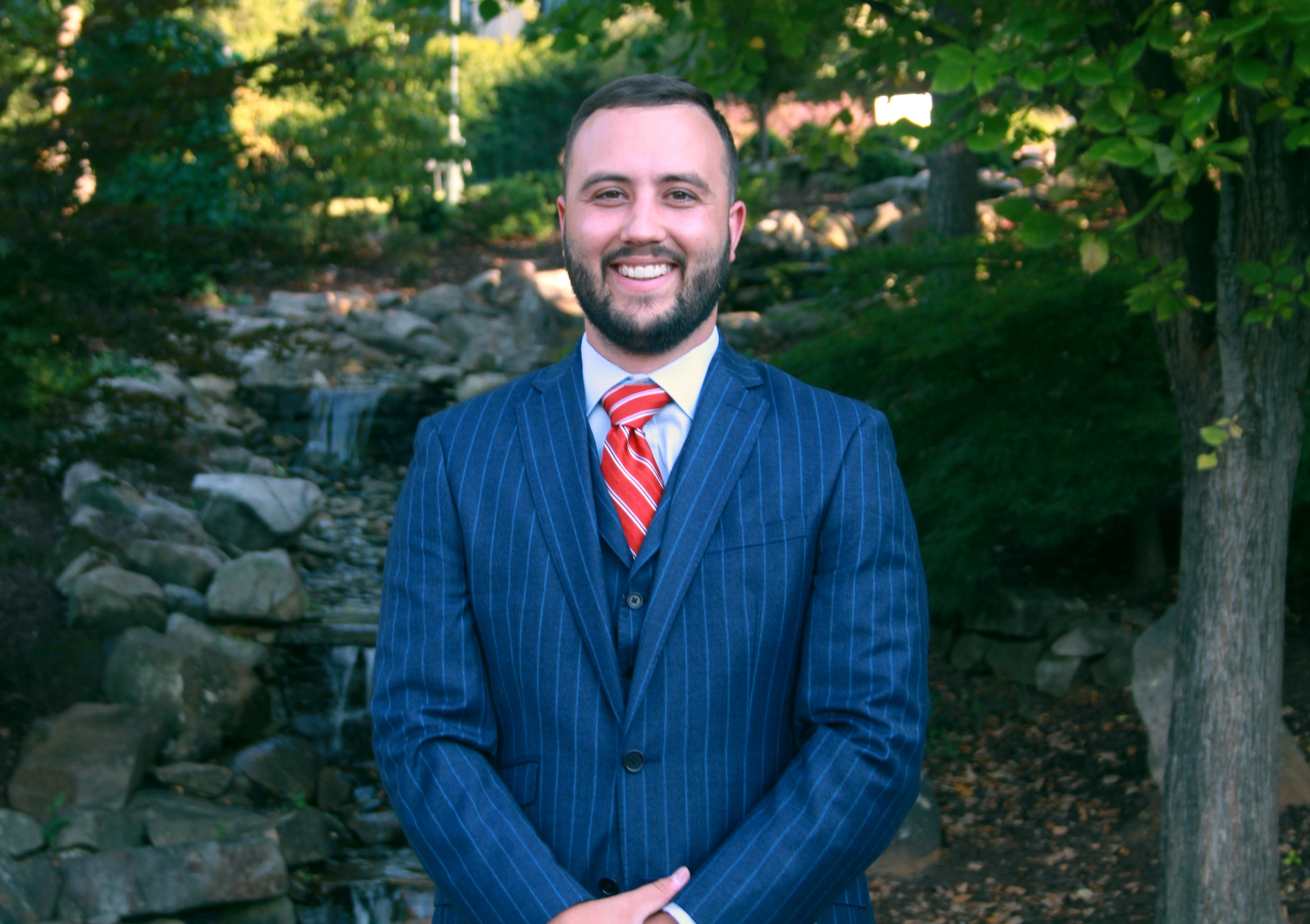 Man in blue pinstripe suit, red tie, smiling, standing outdoors near a small waterfall.