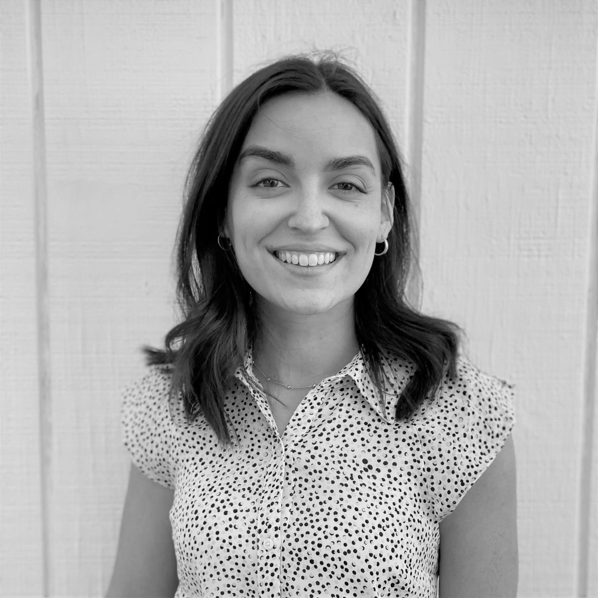 A woman in a polka dot shirt is smiling in front of a white wall.