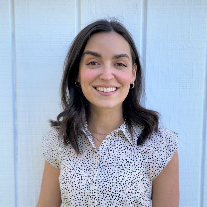 A woman in a polka dot shirt is smiling in front of a white wall.