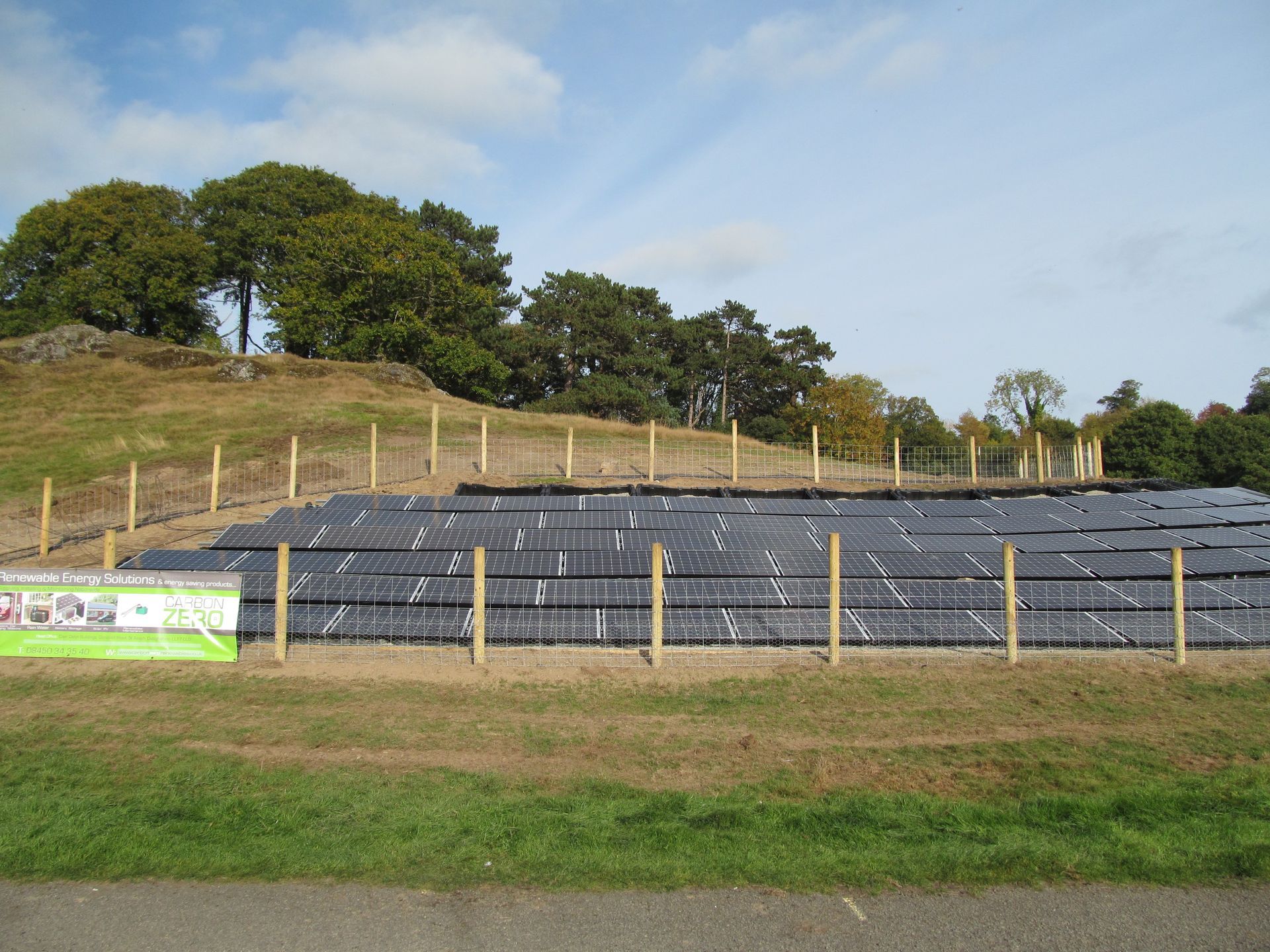 Covered field with black tarp, wooden posts, grassy foreground, and trees on a hillside under a blue sky