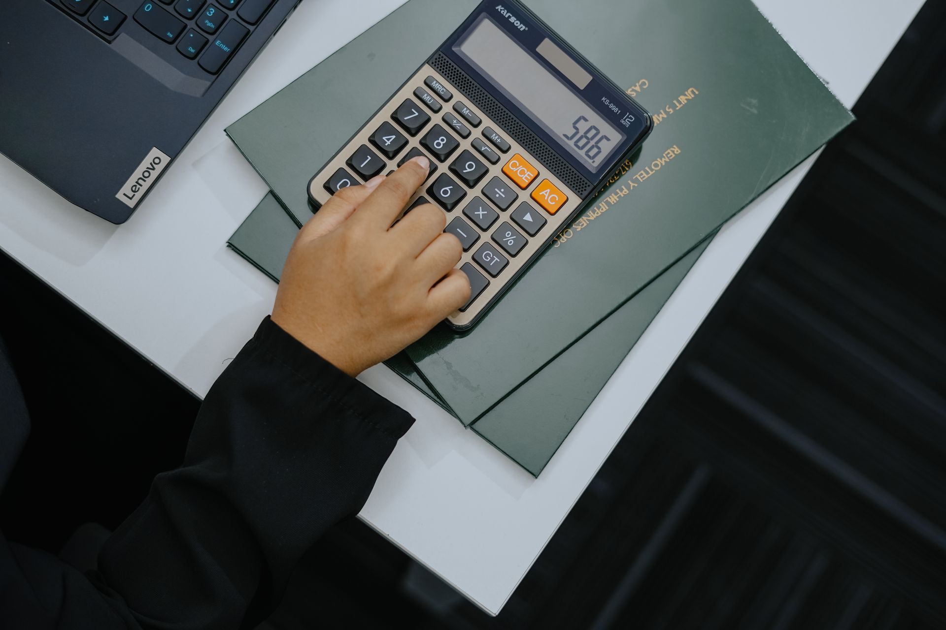 Person using a calculator on a desk, with a laptop and green folder.