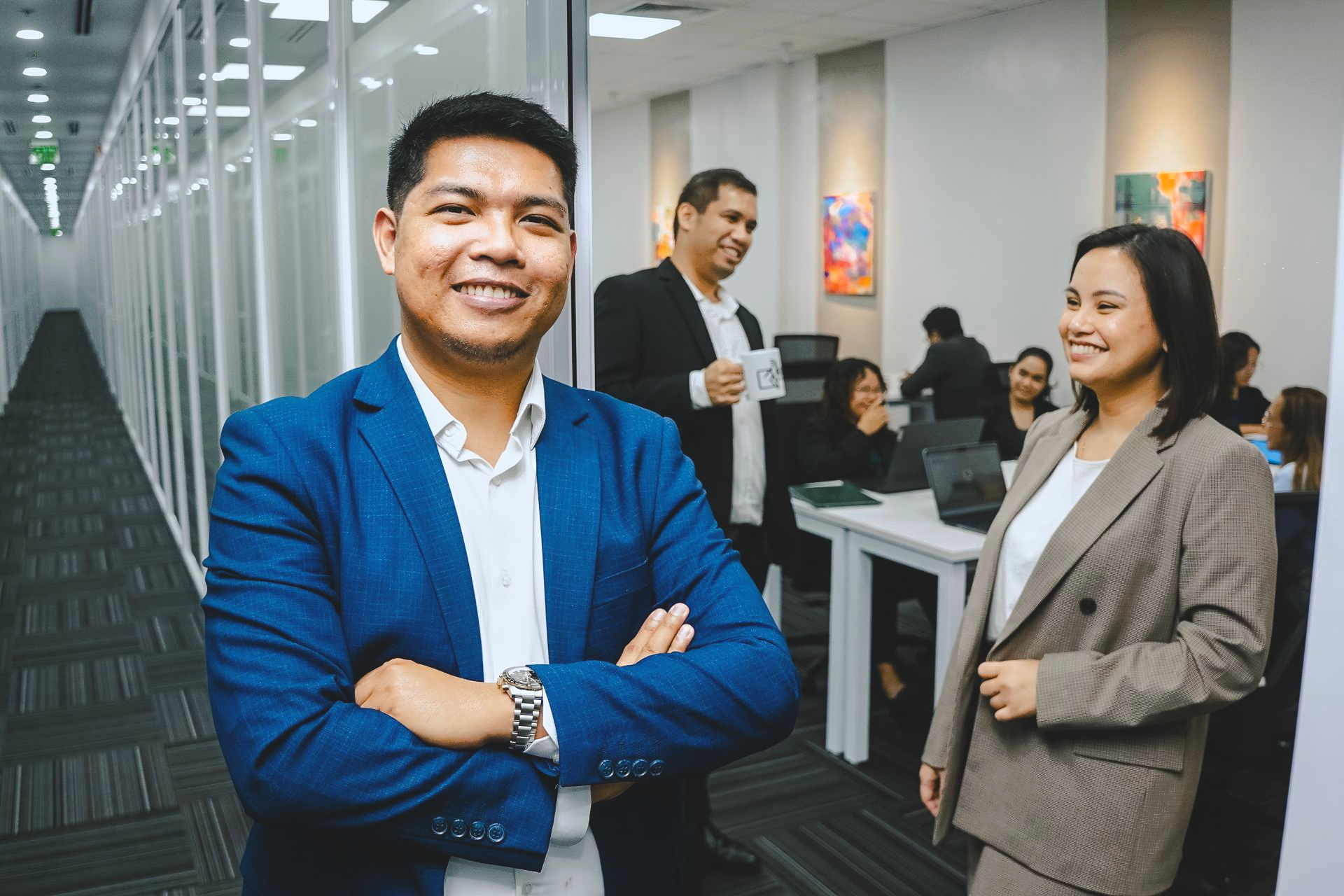 Man in blue blazer smiles, arms crossed, in office. Woman in blazer stands beside him. Other people work in background.