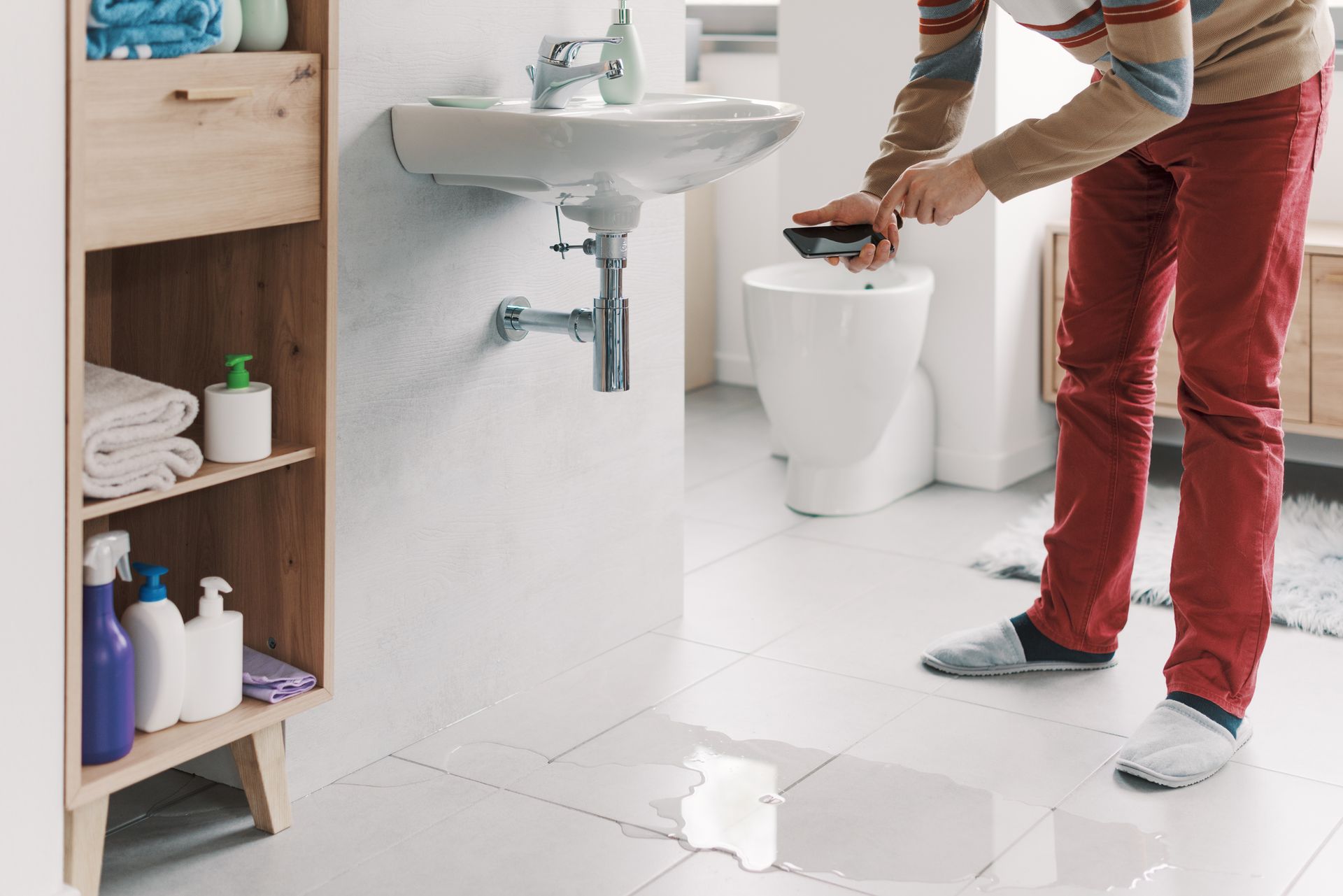 A leaking bathroom sink is spilling water on the floor, and a man is calling a plumber on the phone.