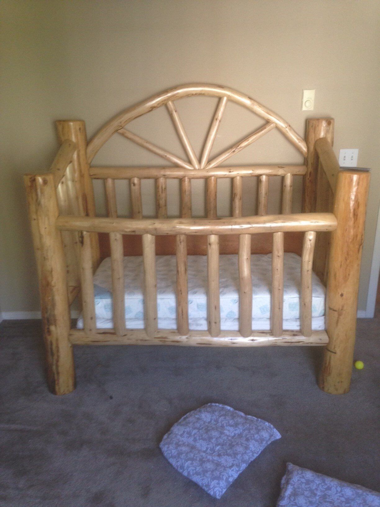 Rustic wooden crib with a mattress, in a room with neutral walls and a carpet.