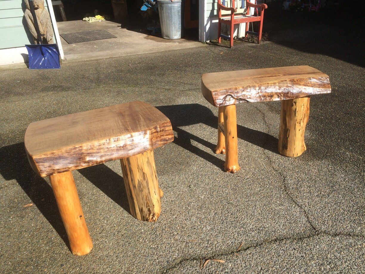 Two rustic wooden stools with natural bark tops and log legs sit outdoors on gravel.
