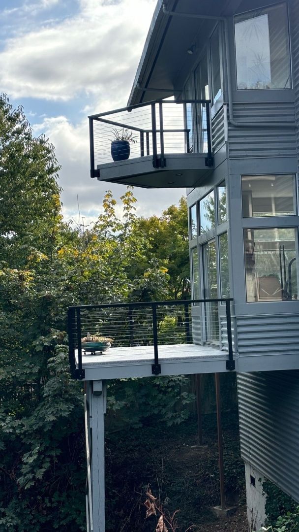 Two-story gray house with cantilevered decks, black railings, and expansive windows. Surrounded by trees.