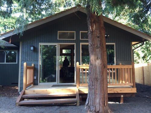 Gray house with a wooden deck and a tree in front. Sliding glass door is open.