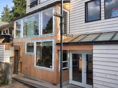 Exterior of a house undergoing construction with large windows, unfinished siding, and a glass-paneled roof.