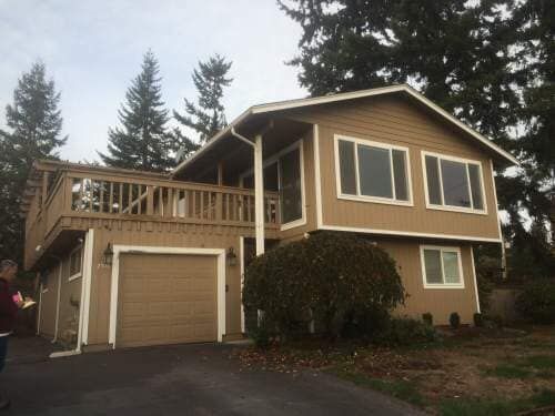 Tan two-story house with a deck, garage, and a driveway, set against a backdrop of trees.